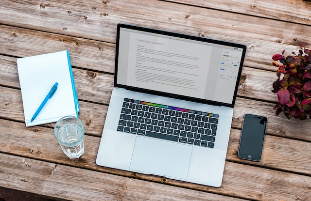 silver macbook beside iphone and clear drinking glass on brown wooden top